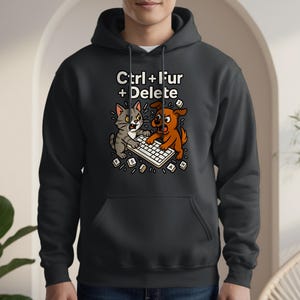 以下が含まれることがあります： 「Ctrl + Fur + Delete」の白い文字が入った、キーボードを巡って戦う漫画の猫と犬が描かれた濃い灰色のパーカー。猫はグレーで、犬は茶色です。パーカーにはフロントポケットとフードが付いています。