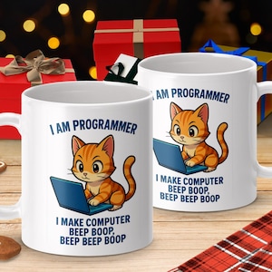 Könnte beinhalten: Zwei weiße Keramikbecher mit einer Cartoon-orangefarbenen Tabby-Katze auf einem blauen Laptop. Die Becher tragen den Text "I AM PROGRAMMER" und "I MAKE COMPUTER BEEP BOOP, BEEP BEEP BOOP". Festlicher Hintergrund mit Geschenkboxen und einem Lebkuchenmann.