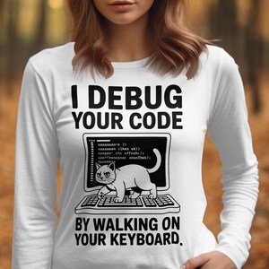 Puede incluir: Camiseta blanca de manga larga con el texto "I DEBUG YOUR CODE" y un gato en un teclado. El texto "BY WALKING ON YOUR KEYBOARD" está debajo. La camiseta es de estilo casual.