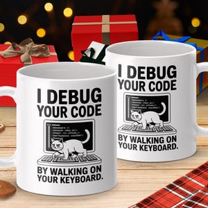 May include: Two white ceramic mugs with the text "I DEBUG YOUR CODE BY WALKING ON YOUR KEYBOARD." Each mug features a graphic of a cat on a keyboard. The background includes wrapped gifts and holiday decorations.