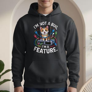 Puede incluir: Una sudadera con capucha gris oscuro con un gráfico de un gato sentado en un servidor, rodeado de cables coloridos. El texto dice "I'M NOT A BUG, I'M A FEATURE." La sudadera tiene un bolsillo delantero y una capucha.
