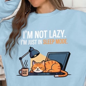 May include: Pale blue sweatshirt featuring the text "I'M NOT LAZY. I'M JUST IN SLEEP MODE." A cartoon cat is sleeping on a laptop next to a mug that says "SLEEP MODE". A desk lamp shines light on the cat.