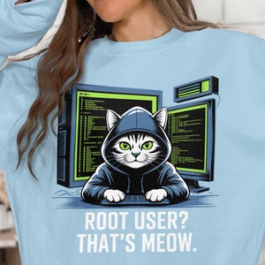 May include: Pale blue sweatshirt with a cartoon cat in a hoodie, positioned before two computer monitors. The cat has green eyes and is using a keyboard. The text on the shirt says "ROOT USER? THAT'S MEOW."