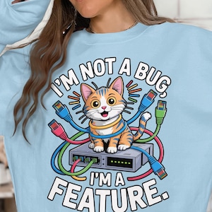 Puede incluir: Sudadera azul claro con una ilustración de gato de dibujos animados. El gato es naranja y blanco, rodeado de cables de red coloridos, y está sentado en un servidor. El texto dice: "I'm not a bug, I'm a feature."