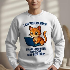 Puede incluir: Sudadera blanca con un gato atigrado naranja de dibujos animados usando una computadora portátil azul. El texto encima del gato dice "I AM PROGRAMMER" y debajo "I MAKE COMPUTER BEEP BOOP, BEEP BEEP BOOP."