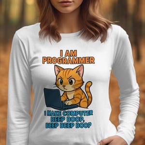 以下が含まれることがあります： 白い長袖シャツ。オレンジ色の縞模様の猫がノートパソコンを使っている漫画のイラストが描かれています。猫の上には「I AM PROGRAMMER」、下には「I MAKE COMPUTER BEEP BOOP, BEEP BEEP BOOP」と書かれています。
