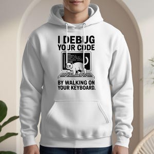May include: White hoodie with a front graphic. The graphic features a laptop with code and a cat walking on the keyboard. Text reads "I debug your code by walking on your keyboard."