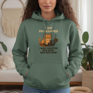 May include: Green hoodie featuring a cat graphic wearing glasses and working on a laptop. The text on the hoodie reads "I AM PROGRAMMER I MAKE COMPUTER BEEP BOOP, BEEP BEEP BOOP."