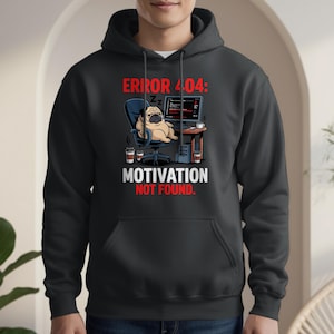 May include: Black hoodie featuring a graphic of a sleeping pug in an office chair in front of a computer. The text reads "ERROR 404: MOTIVATION NOT FOUND." Two coffee cups are on the desk. The hoodie has a front pocket and a drawstring hood.