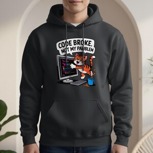 Puede incluir: Sudadera con capucha negra con un diseño de gato de dibujos animados. El gato es naranja y blanco, con un ordenador y el texto "CODE BROKE. NOT MY PAWBLEM." También se muestra una taza azul con café derramado.