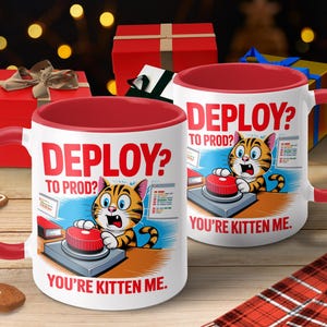 May include: White ceramic mug with a red interior and handle. The mug features a cartoon cat pushing a red button with the text "DEPLOY? TO PROD? YOU'RE KITTEN ME." The background includes gift boxes and a gingerbread cookie.