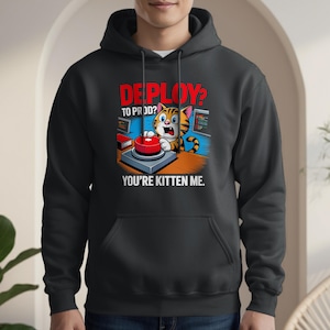 Könnte beinhalten: Schwarzer Kapuzenpullover mit einem Cartoon-Katzendesign. Die Katze drückt einen roten Knopf, darüber steht "DEPLOY? TO PROD? YOU'RE KITTEN ME." Die Katze ist orange und braun. Das Design beinhaltet Computerbildschirme.