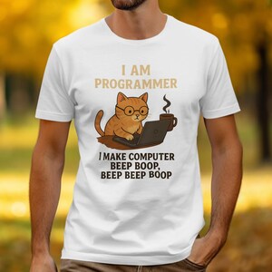 以下が含まれることがあります： メガネをかけた猫がノートパソコンで作業している漫画のグラフィックが描かれた白いTシャツ。「I AM PROGRAMMER」と猫の上に書かれ、その下に「I MAKE COMPUTER BEEP BOOP, BEEP BEEP BOOP」と書かれています。コーヒーカップも画像にあります。