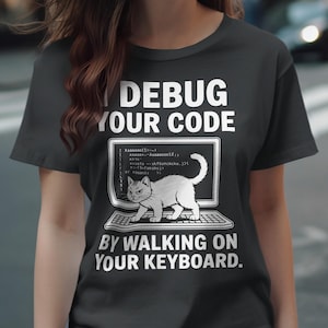 May include: A dark grey t-shirt featuring the text "I DEBUG YOUR CODE BY WALKING ON YOUR KEYBOARD." The design shows a computer screen with code and a white cat on a keyboard.