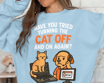 Op de afbeelding: Lichtblauw sweatshirt met de tekst "HAVE YOU TRIED TURNING THE CAT OFF AND ON AGAIN?". De afbeelding toont een cartoonkat op een kapotte laptop en een hond met een headset die naar een computerscherm met een foutmelding kijkt.