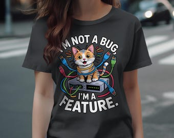 Può includere: T-shirt grigio scuro con un gatto dei cartoni animati a strisce arancioni e bianche seduto su un dispositivo di rete. Il testo recita "I'M NOT A BUG, I'M A FEATURE." Cavi di rete colorati circondano il gatto e il dispositivo.