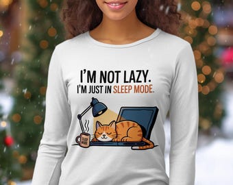 Op de afbeelding: Wit shirt met lange mouwen met de tekst "I'M NOT LAZY. I'M JUST IN SLEEP MODE." Een cartoonkat slaapt op een laptop. Een mok met de woorden "SLEEP MODE" staat naast de laptop. Een bureaulamp is verlicht.