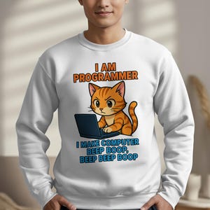 Puede incluir: Sudadera blanca con el texto "I AM PROGRAMMER" en naranja y azul. Un gato atigrado naranja de dibujos animados está sentado frente a una computadora portátil. Debajo está el texto "I MAKE COMPUTER BEEP BOOP, BEEP BEEP BOOP".