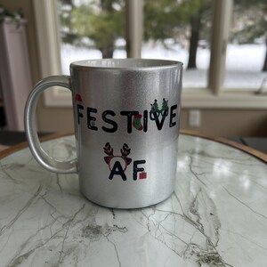May include: Silver glitter mug with the text "FESTIVE AF" in black. The mug features festive holiday graphics, including a Santa hat, Christmas tree, and reindeer antlers. The mug is on a marble-patterned surface.