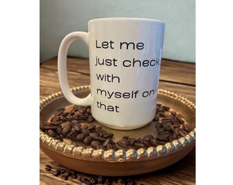 以下が含まれることがあります： 「Let me just check with myself on that」の黒い文字が入った白いセラミックコーヒーマグ。マグカップは、コーヒー豆に囲まれた金色のトレイの上に置かれています。マグカップには大きなハンドルが付いています。
