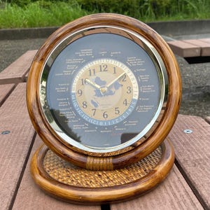 May include: A circular wooden world time clock with a gold face and a dark background. The clock has a gold bezel and a woven base. The clock face displays the time and various world cities. The clock measures approximately 15 cm in diameter.