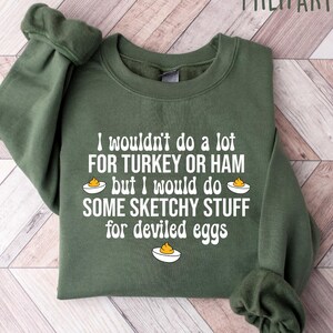 May include: Military green sweatshirt with white text: "I wouldn't do a lot FOR TURKEY OR HAM but I would do SOME SKETCHY STUFF for deviled eggs." The design includes illustrations of deviled eggs. The sweatshirt is folded on a light wood surface.