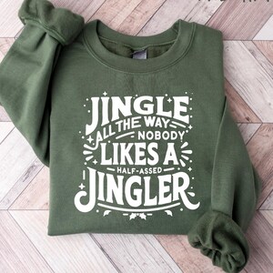 May include: A military green sweatshirt with the text "Jingle All The Way Nobody Likes A Half-Assed Jingler" in white. The sweatshirt is folded on a light wood-grain surface.