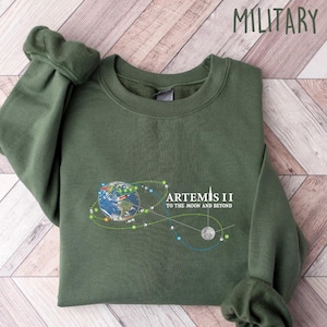 May include: Military green sweatshirt with the text "ARTEMIS II TO THE MOON AND BEYOND" and a graphic of the Earth, moon, and connecting lines. The sweatshirt is folded on a light wood-grain surface.