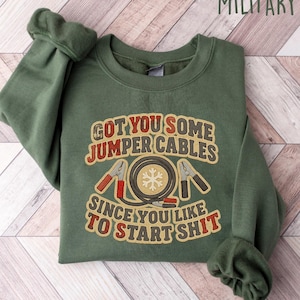 May include: Military green sweatshirt with the text "GOT YOU SOME JUMPER CABLES SINCE YOU LIKE TO START SHIT". The graphic includes jumper cables and a snowflake design. The sweatshirt is laid flat on a light wood grain surface.