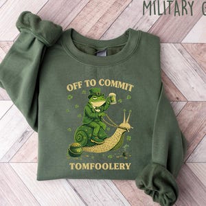 May include: Military green sweatshirt with a St. Patrick's Day graphic. The design features a frog in a leprechaun outfit riding a snail, holding a beer, with the text "OFF TO COMMIT TOMFOOLERY". The sweatshirt is made of a soft material.