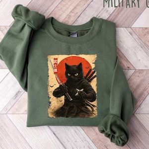 May include: Military green sweatshirt featuring a graphic of a black ninja cat with a sword and throwing star against a red sun. The cat is wearing a ninja outfit. The text "Military Green" is at the top.