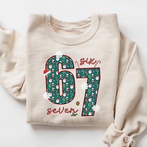 May include: A beige sweatshirt with the numbers "67" in a Christmas-themed pattern. The numbers are filled with Santa Claus, snowflakes, and holly. The word "Six" and "Seven" are written above and below the numbers.
