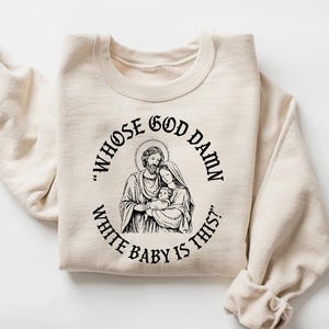 May include: A sand-colored sweatshirt with a black graphic of a religious family and the text "WHOSE GOD DAMN WHITE BABY IS THIS?" The sweatshirt is folded with the sleeves rolled up.