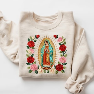 May include: A sand-colored sweatshirt featuring an embroidered image of the Virgin of Guadalupe, surrounded by red and pink roses. The design includes a gold halo and a detailed depiction of the Virgin Mary. The sleeves are rolled up.