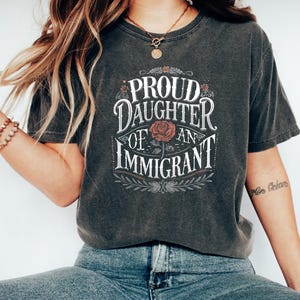 May include: Dark gray t-shirt with the text "PROUD DAUGHTER OF AN IMMIGRANT" in a decorative font, featuring a rose graphic. The shirt is paired with blue jeans and white sneakers.