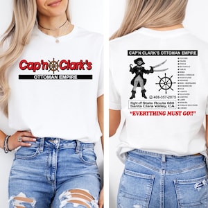 May include: A white t-shirt featuring the red and black text Cap'n Clark's OTTOMAN EMPIRE on the front. The back displays a graphic of a pirate, a ship wheel, a list of furniture items, a phone number, an address, and the text EVERYTHING MUST GO.