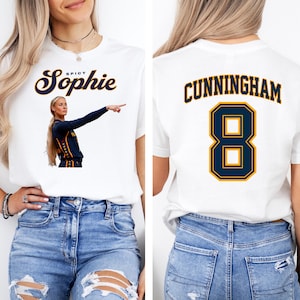 May include: A white short-sleeve t-shirt featuring a graphic of a basketball player pointing with the text Spicy Sophie on the front and the name CUNNINGHAM with the number 8 in dark blue and yellow on the back, paired with blue denim jeans.