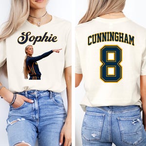 May include: A cream-colored short-sleeve t-shirt. The front features the name Sophie in navy blue script above a graphic of a basketball player. The back displays the name CUNNINGHAM in navy blue block letters above the large navy blue number 8.