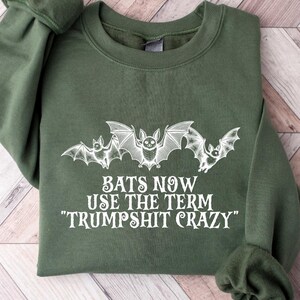 May include: Military green sweatshirt with white bat graphic and the text "BATS NOW USE THE TERM 'TRUMPSHIT CRAZY'". The long-sleeved top is folded on a light wood-grain surface.