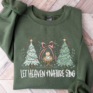 May include: Military green sweatshirt with a Christmas-themed graphic. The design includes two Christmas trees, a baby in a manger, and the text "LET HEAVEN & NATURE SING" in white. The sweatshirt is folded on a light wood-grain surface.
