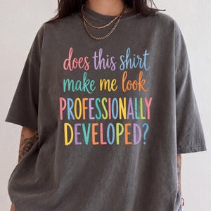 May include: A charcoal gray short-sleeve t-shirt featuring the multicolored text "does this shirt make me look PROFESSIONALLY DEVELOPED?" printed on the front. The shirt has a crew neckline and a relaxed fit, showcasing a casual style.