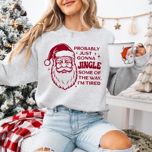May include: A light gray sweatshirt with a red Santa Claus graphic and the text "Probably Just Gonna Jingle Some of the Way, I'm Tired." A white mug with a Santa Claus graphic is held in the hand.