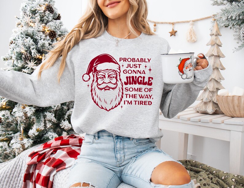 May include: A light gray sweatshirt with a red Santa Claus graphic and the text "Probably Just Gonna Jingle Some of the Way, I'm Tired." A white mug with a Santa Claus graphic is held in the hand.