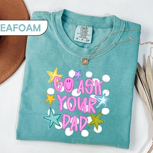 May include: Seafoam green t-shirt with the phrase "GO ASK YOUR DAD" in pink, with star and dot graphics. The shirt has a crew neck and short sleeves. A gold necklace with a pendant is draped on the shirt.