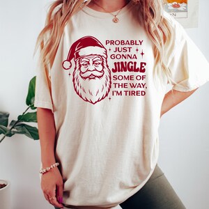 May include: Cream-colored t-shirt featuring a Santa Claus graphic in red with the text "Probably Just Gonna Jingle Some of the Way, I'm Tired." The Santa graphic includes a hat, beard, and a stern expression. The shirt is a casual, festive garment.