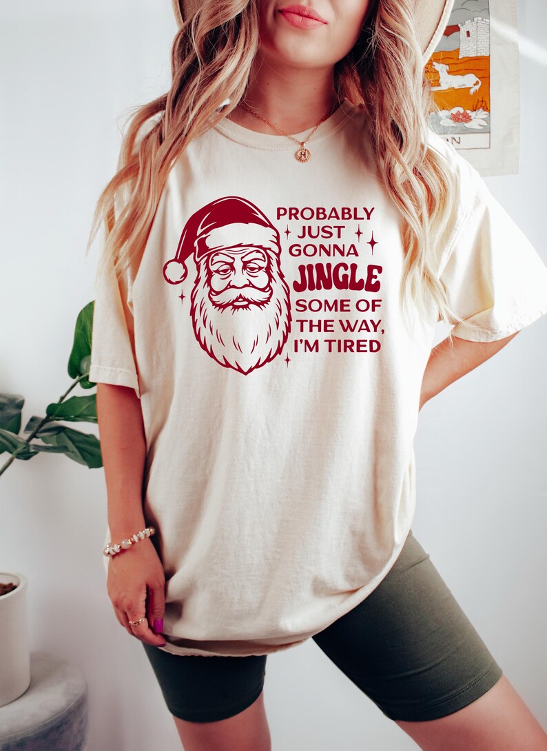 May include: Cream-colored t-shirt featuring a Santa Claus graphic in red with the text "Probably Just Gonna Jingle Some of the Way, I'm Tired." The Santa graphic includes a hat, beard, and a stern expression. The shirt is a casual, festive garment.