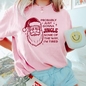 May include: A light pink sweatshirt with a Santa Claus graphic and the text "PROBABLY JUST GONNA JINGLE SOME OF THE WAY, I'M TIRED." The sweatshirt is paired with light blue shorts. A mug with a smiley face is held in the hand.