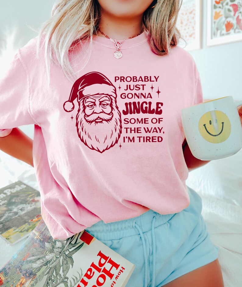 May include: A light pink sweatshirt with a Santa Claus graphic and the text "PROBABLY JUST GONNA JINGLE SOME OF THE WAY, I'M TIRED." The sweatshirt is paired with light blue shorts. A mug with a smiley face is held in the hand.