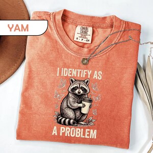 May include: A yam-colored t-shirt featuring a graphic of a raccoon holding a coffee cup surrounded by butterflies. The text reads I IDENTIFY AS A PROBLEM. The shirt has a crew neckline and a Comfort Colors brand label visible at the inner collar.