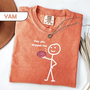 May include: A terracotta-colored t-shirt featuring a white stick figure graphic holding a pink brain illustration. The text "Hey you dropped this" is printed in white above the brain. The shirt has a Comfort Colors label showing size S-CH.